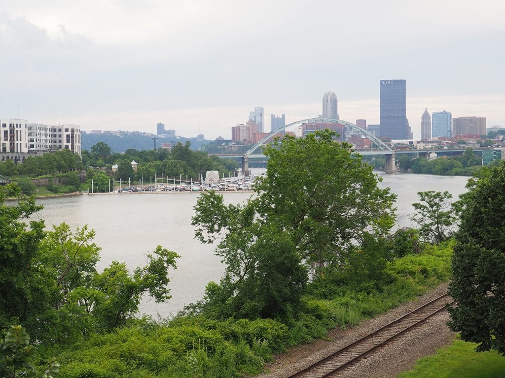 a photo of a river with a railroad amongst greenery on its near bank, and a city skyline in the background