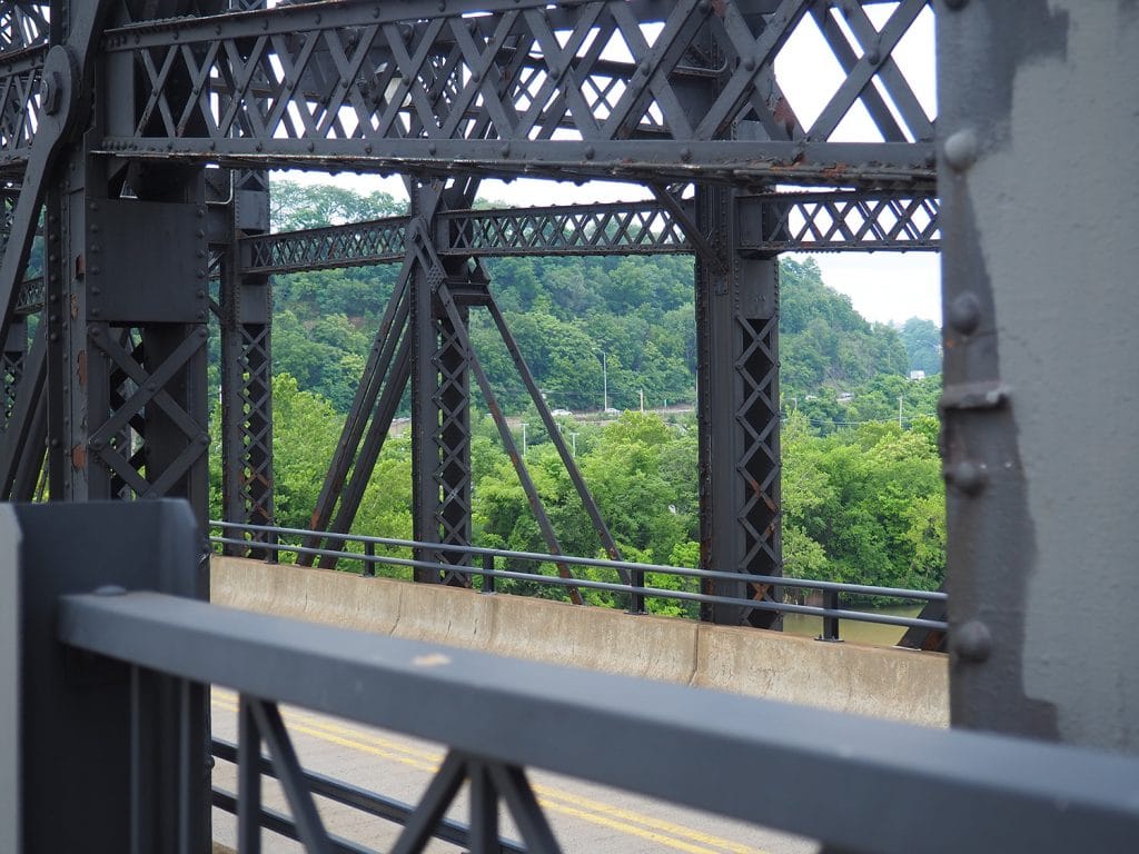 a photo of forested hills framed by the beams of a grey metal bridge in the foreground