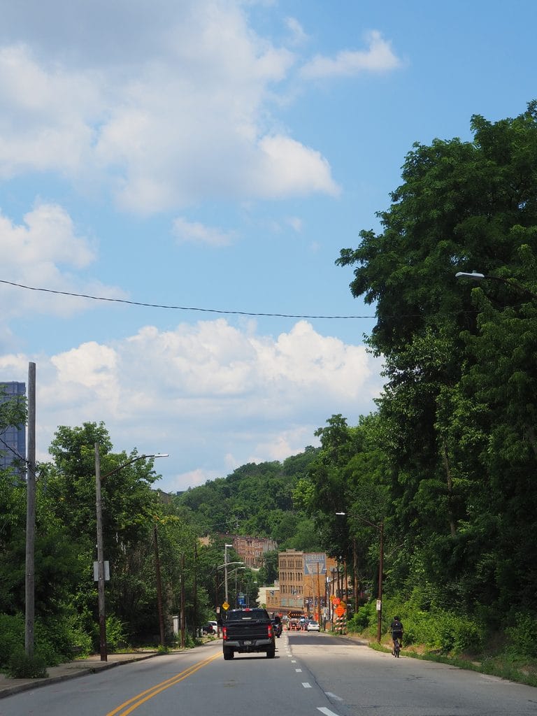 a photo of a downhill street, surrounded by tall trees and a blue cloudy sky above