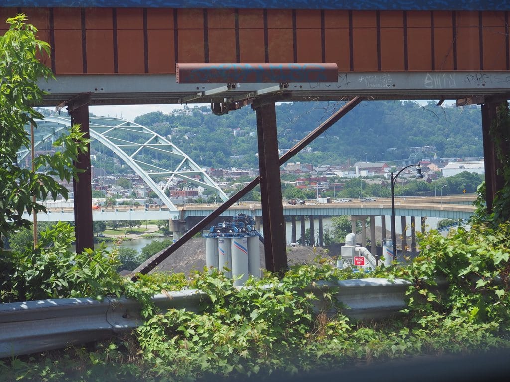 a photo of an arch bridge over a river, framed by greenery and industrial elements in the foreground